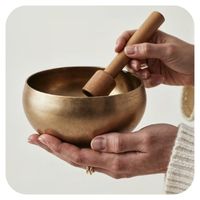 Hands holding a bronze singing bowl with a wooden mallet on a light background